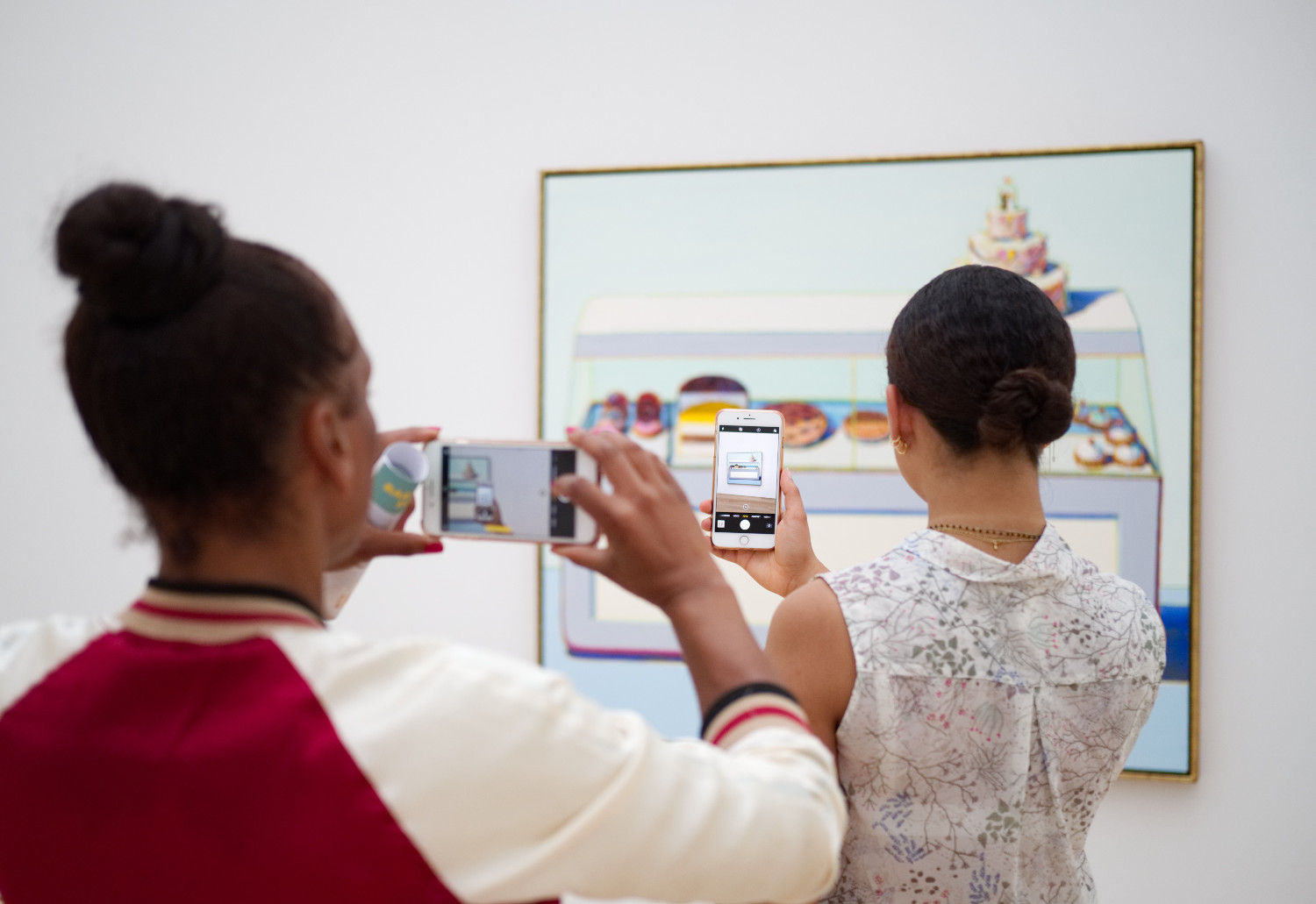 © Image: Christian Fregna Two people have their backs to the camera as they both take a photo of a painting hanging on the wall. The person in the back is taking her photo of the painting through the phone screen of the person of the person standing in front of her.