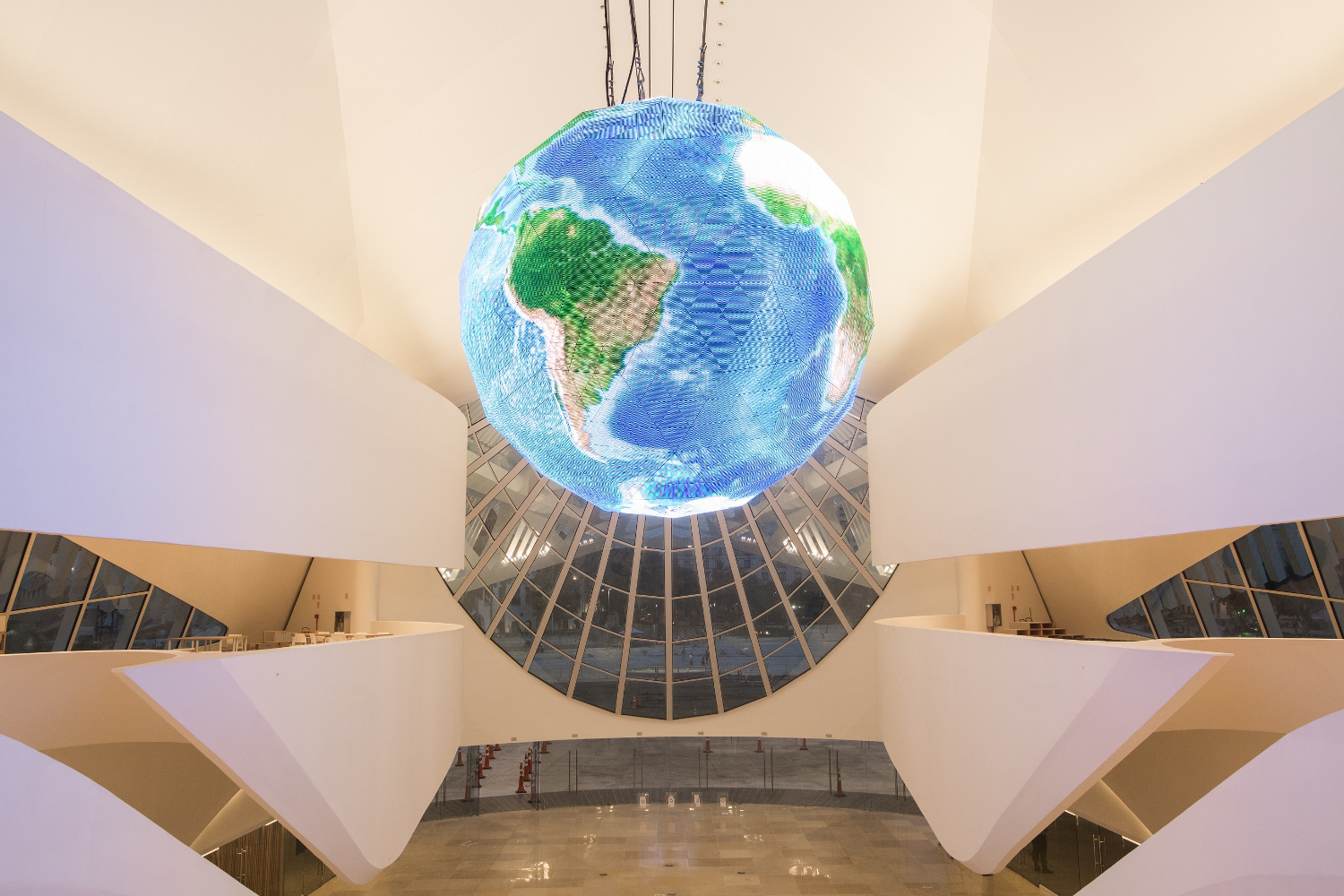 Photo shows a very modern and white building from inside. A big, illuminated globe is hanging from the ceiling.