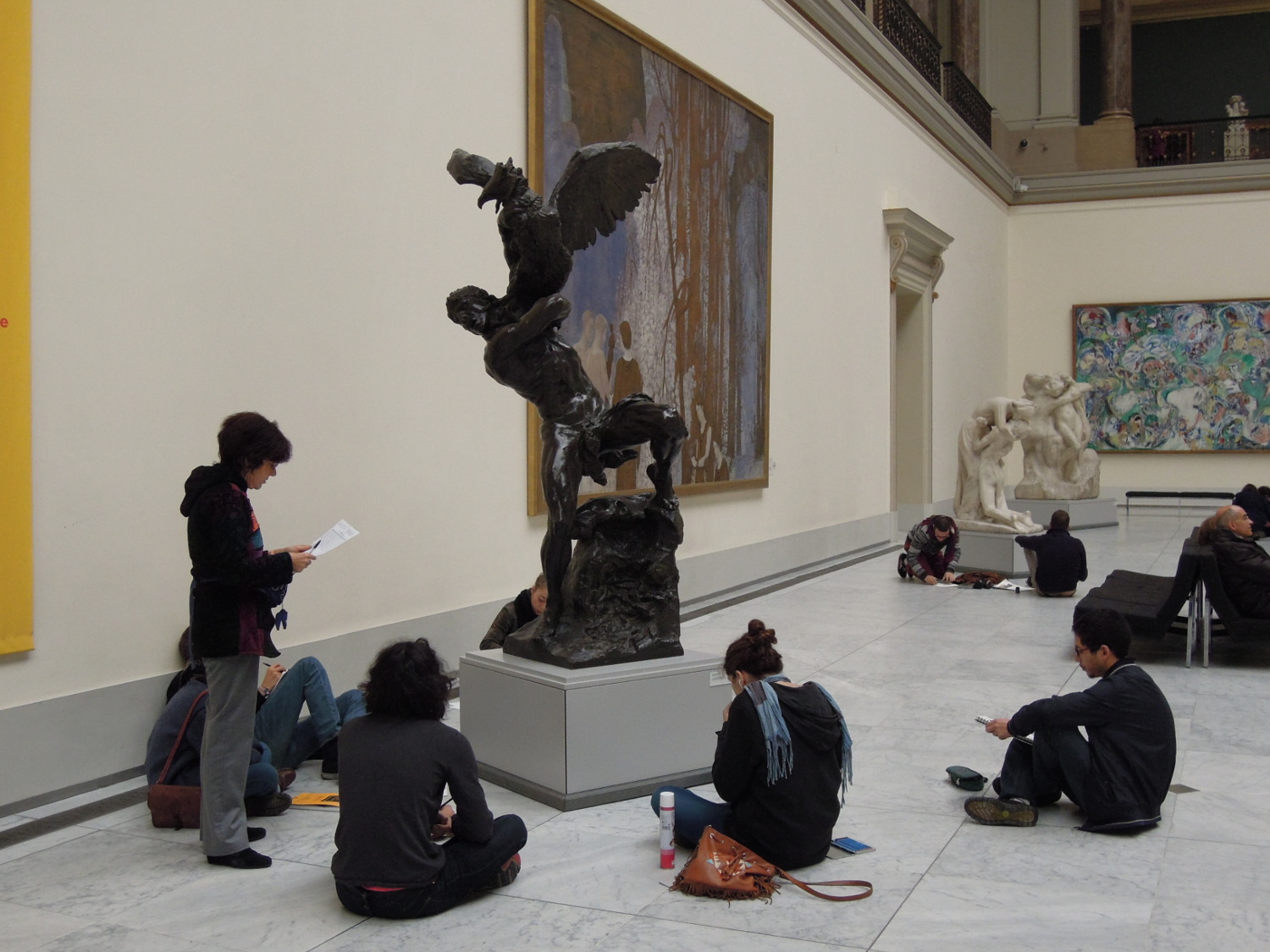  A group of young people is sitting around a sculpture in a museum space. Another person is standing and holding a piece of paper.