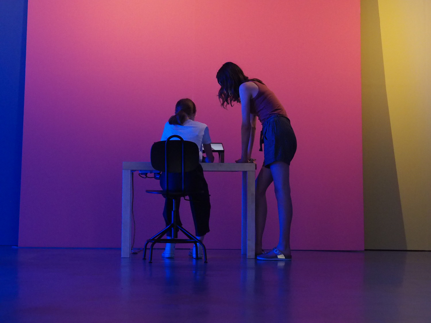  Two children are photographed from the back in a blue lit exhibition space. One of them is sitting, the other one is standing in front of a tablet.