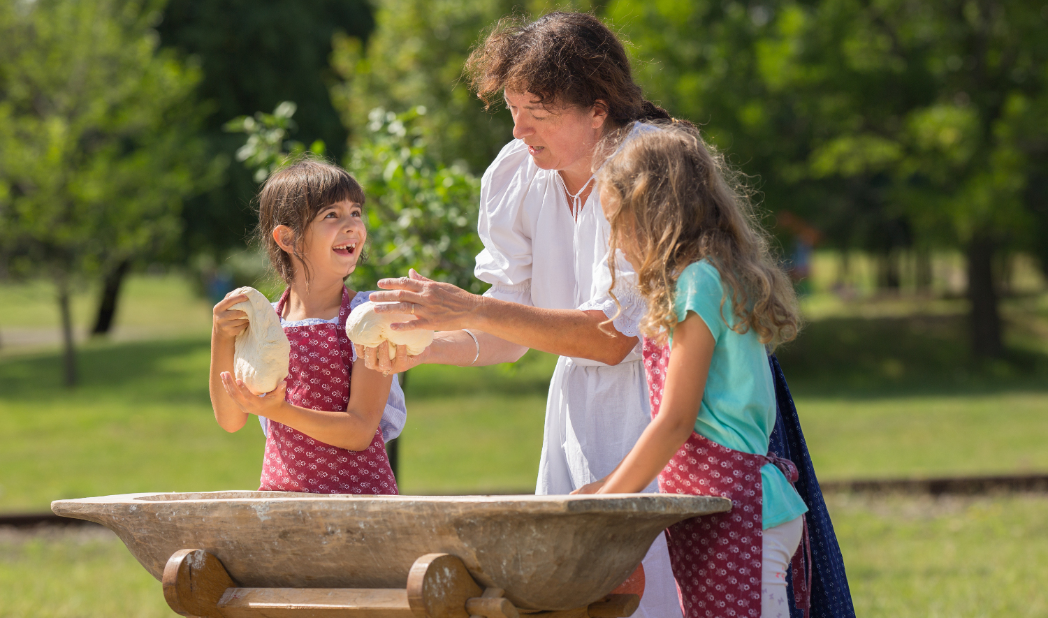 © Hungarian Open Air Museum This photograph depicts a person showing to children how to handle dough. They stand outside behind a wooden trough.