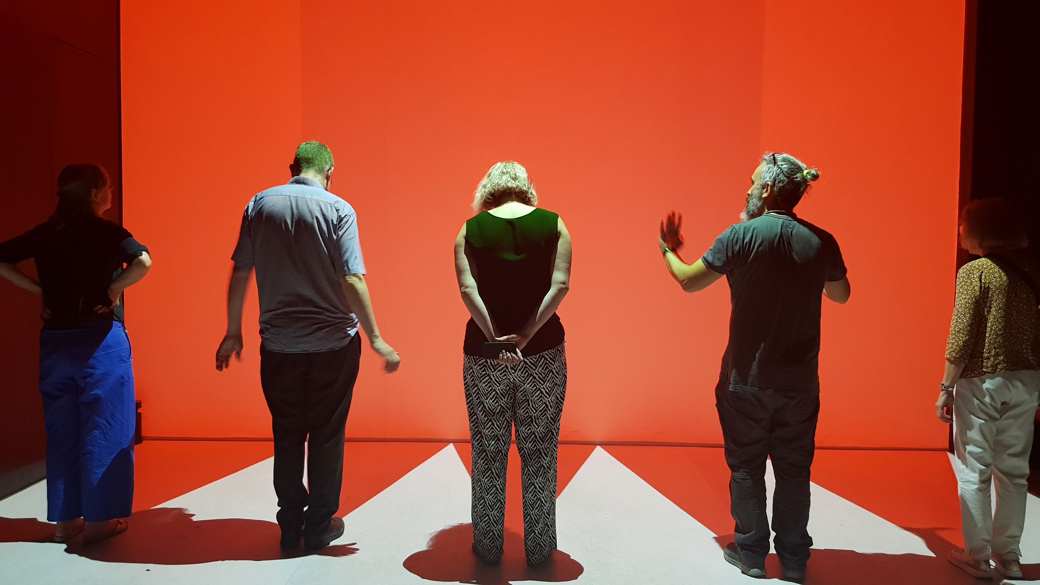 Five people with their back to the camera are standing in front of a big orange wall.  