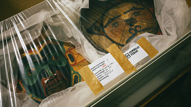 This photo shows two ancient art objects packaged inside a box with foil on top. On the foil is a sticker of the "Humboldt Forum" as well as an inventory sticker.  © Berlin State Museums, Image: Daniel Hofer