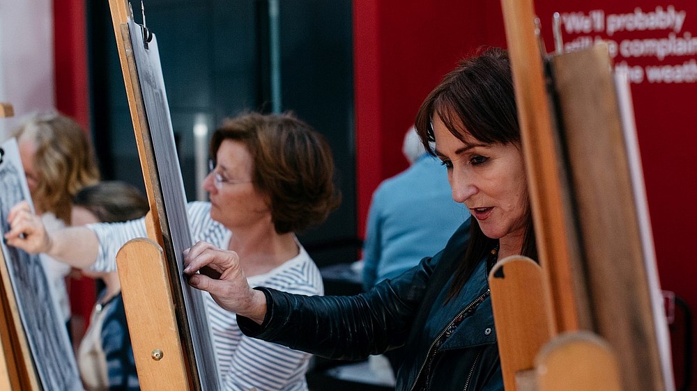 Two people are photographed close up, while drawing with coal on a piece of paper attached to an easel.   