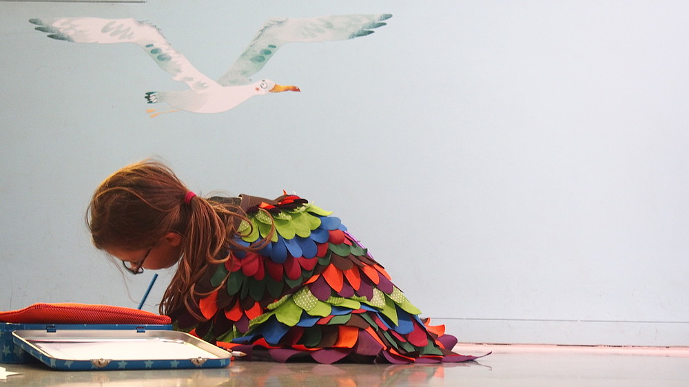 A child dressed in a colourful, eccentric robe is sitting on the floor writing.  