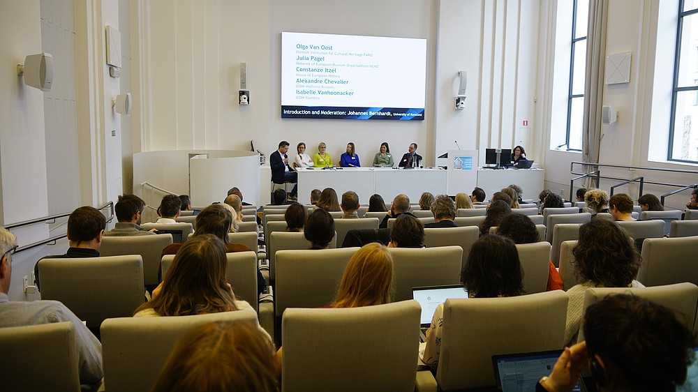 Seated conference participants are seen from the back looking at 7 persons sitting on the stage behind a table. One of them speaks into a hand-held microphone.   © Image: Peter Vand der Plaetsen
