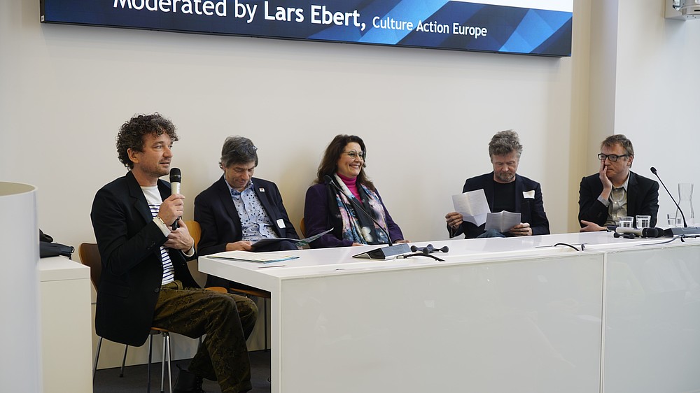 5 persons sit behind a table. Some are looking through notes, one is speaking into a hand-held microphone.  © Image: Peter Vand der Plaetsen