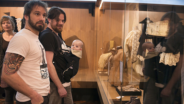  Two men stand next to each other in front of a glass monter displaying old fashioned clothes. One of the men is carrying a baby in a harness on his belly.