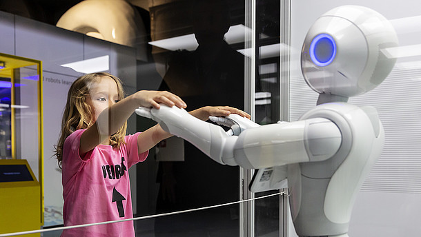  Girl reached over a see through fence to touch the hands of a robot. 