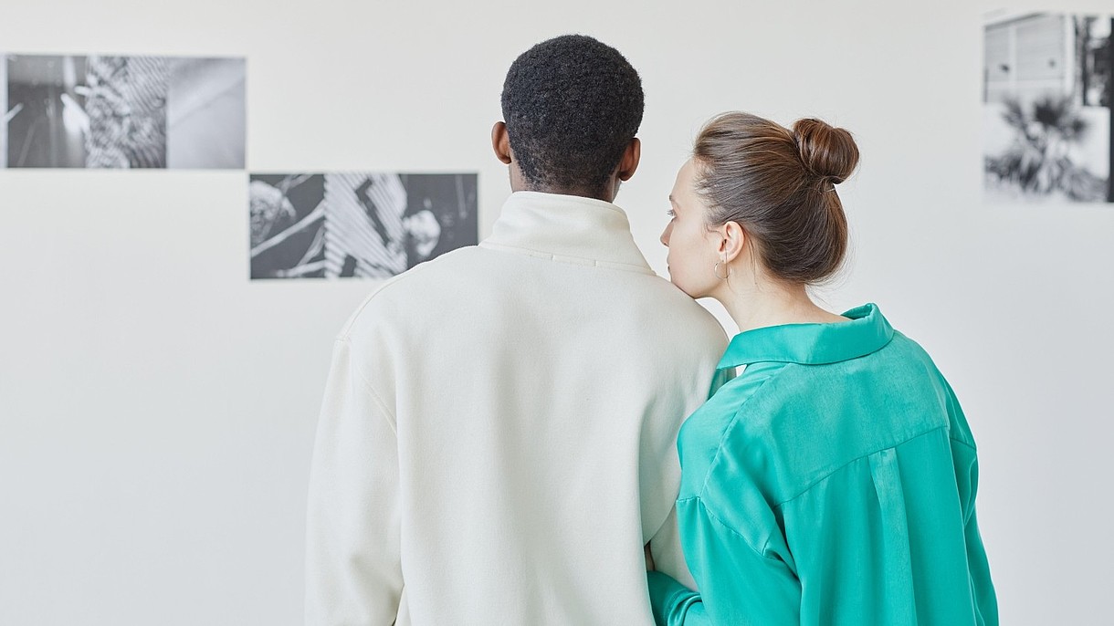 © SeventyFour Images / Alamy Stock Foto © SeventyFour Images / Alamy Stock Foto Two people are standing in a gallery looking at black and white photographs on the wall. One of them is resting their head on the others shoulder.