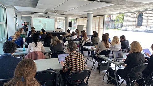 About 40 people are sitting at tables lined up in two rows taking notes and listening to a person who is presenting next to a projector screen.   