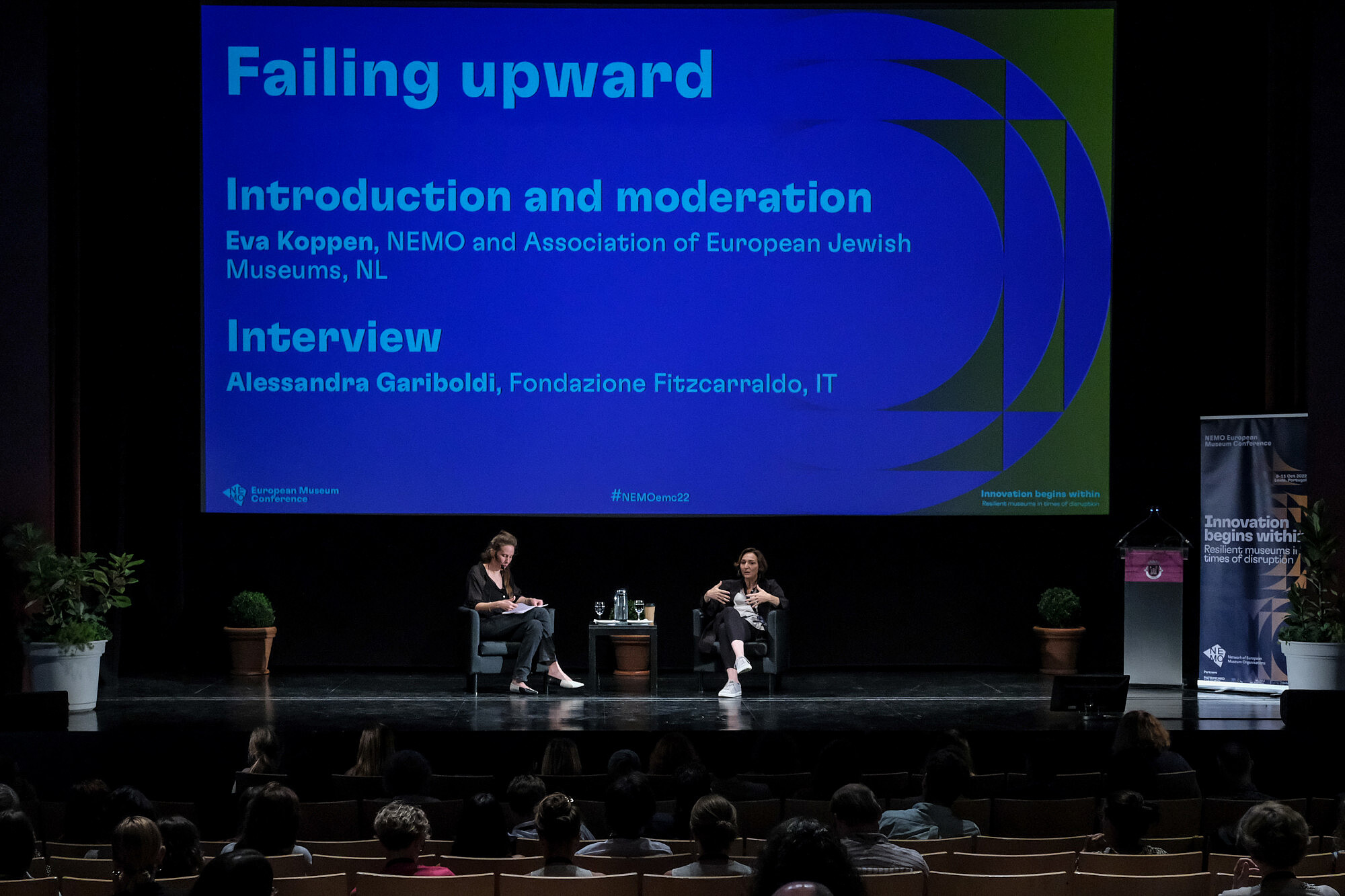 Two people sit on a stage. On of them is gesturing and talking to an audience.  © Image: Jorge Gomes