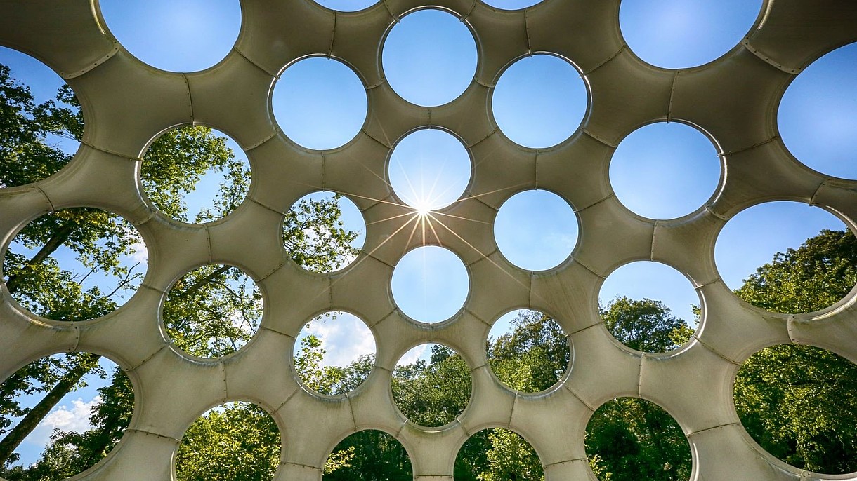  View upwards and through a roof structure with big circular holes. Behind blue skies and trees are visible.