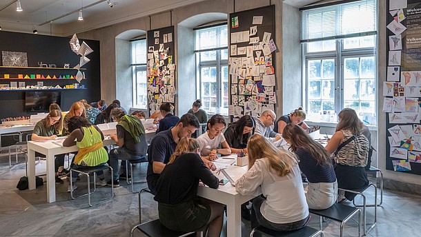  People sit at tables and draw on paper. The walls of the room are covered with colourful drawings.