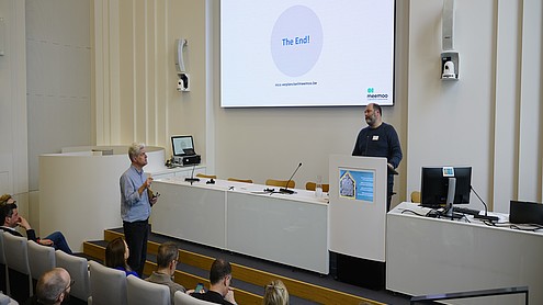 A person stands behind a podium and next to a projected image that reads "the end". He looks at another person who is saying something while standing in front of a seated audience.  © Image: Peter Vand der Plaetsen