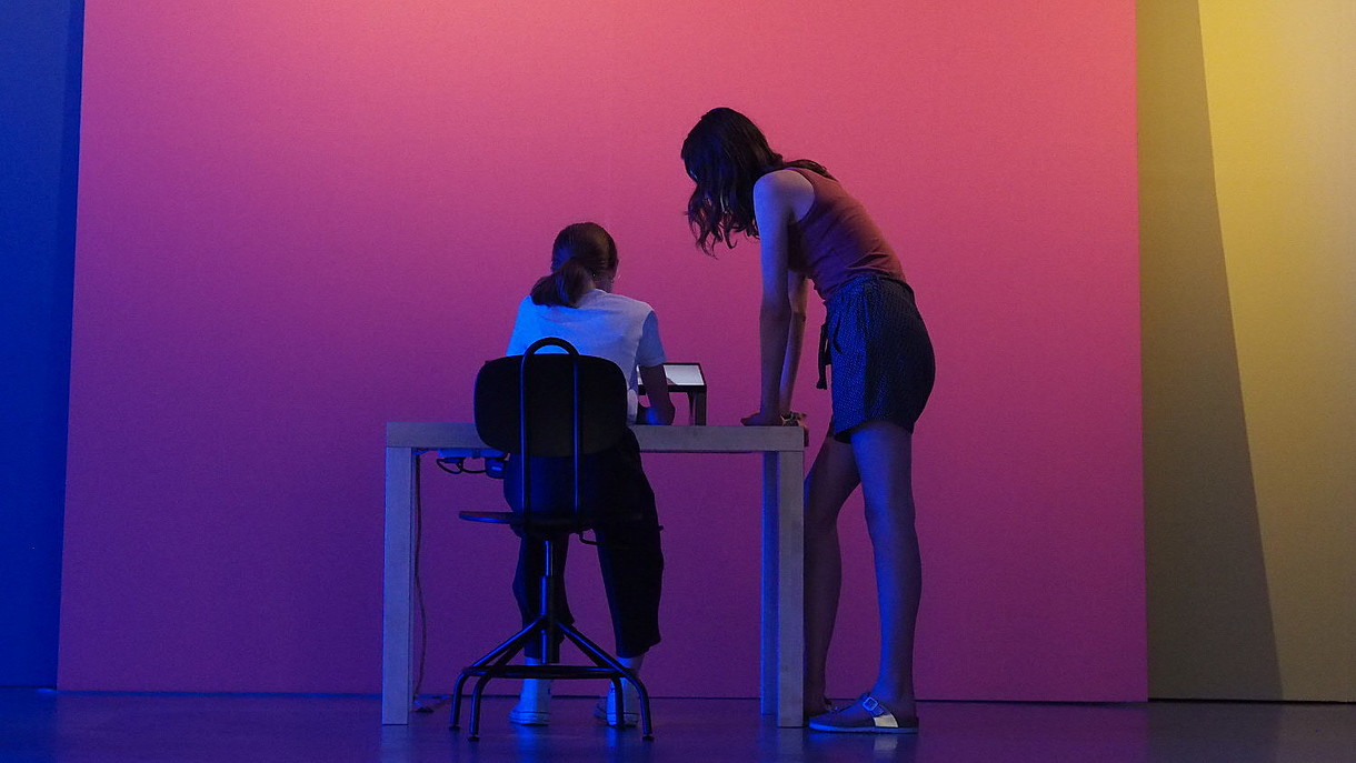  Two children are photographed from the back in a blue lit exhibition space. One of them is sitting, the other one is standing in front of a tablet.