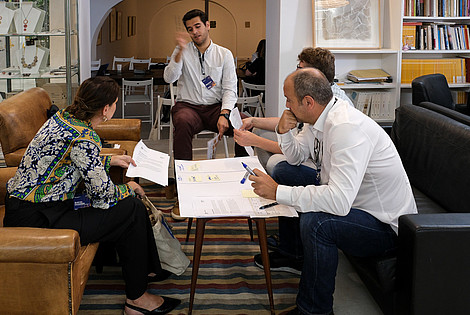 Four people are sitting around a small table. On the table is a large piece of paper with notes. One person is talking an gesturing with their arms, while two of the others are looking at them.  © Image: Jorge Gomes
