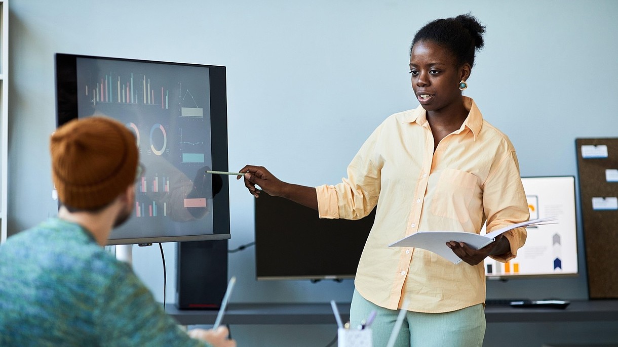 © SeventyFour Images / Alamy Stock Foto A person stands next to a screen and gives a presentation to another person, who sits at a desk.