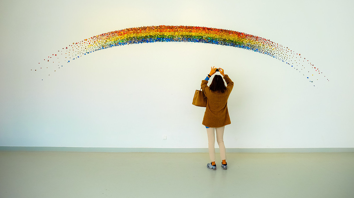 © Image: Francesco Ungaro A person reaches up to take a photo with their phone of a rainbow that is painted on a wall. 