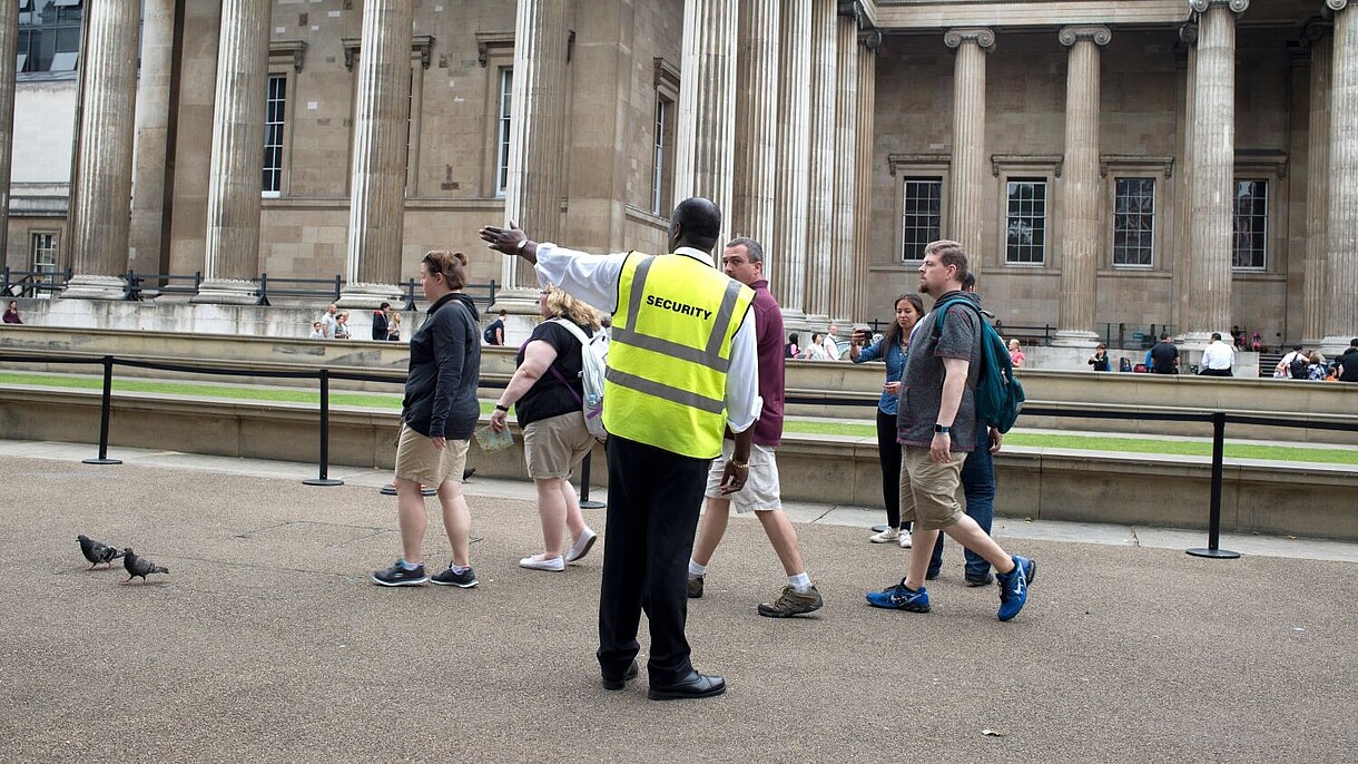 © Jenny Matthews / Alamy Stock Foto A security officer guides a group of people towards the left hand side of the photograph. In the background, people queing in front of a museum can be seen.