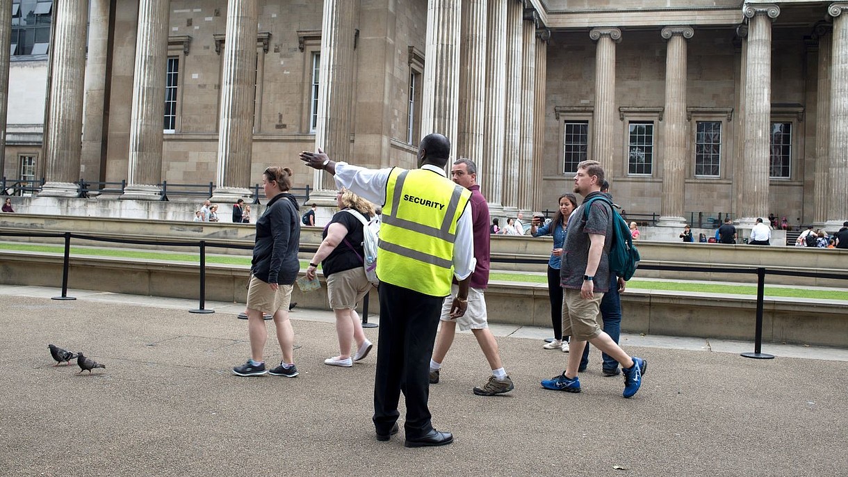 © Jenny Matthews / Alamy Stock Foto A security officer guides a group of people towards the left hand side of the photograph. In the background, people queing in front of a museum can be seen.