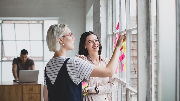 Two people stand in front of a window and talk. They have put different coloured sticky notes on the window. In the background is a person working in front of a laptop.
