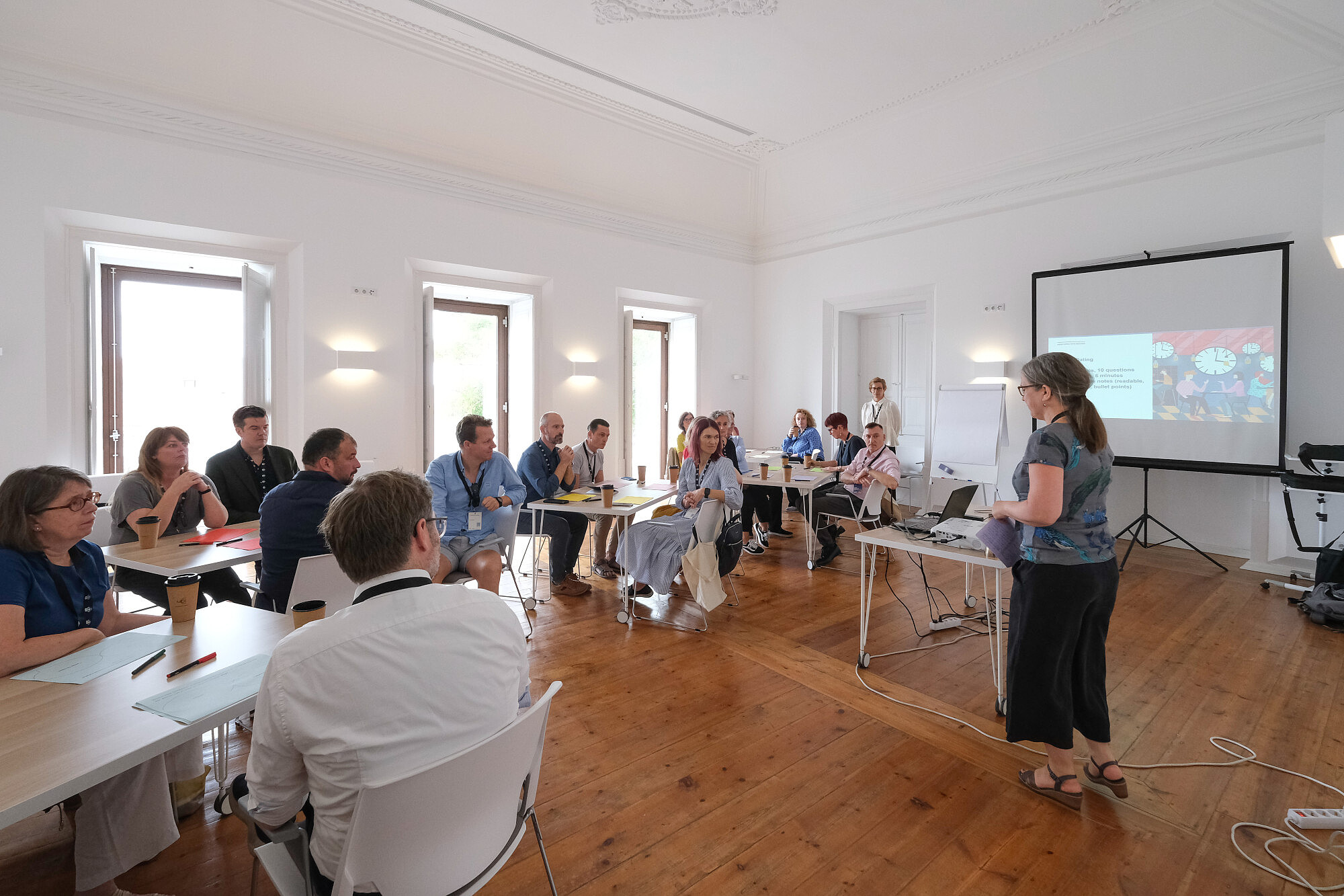 People are sitting on small tables in a conference room. One person is standing and talking. Presentation slides are projected to a screen at the front of the room.  © Image: Jorge Gomes