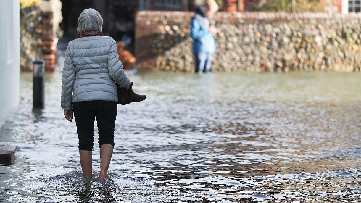 © PA Images / Alamy Stock Foto A person, photographed from the back, is walking barefoot through a flooded street. 