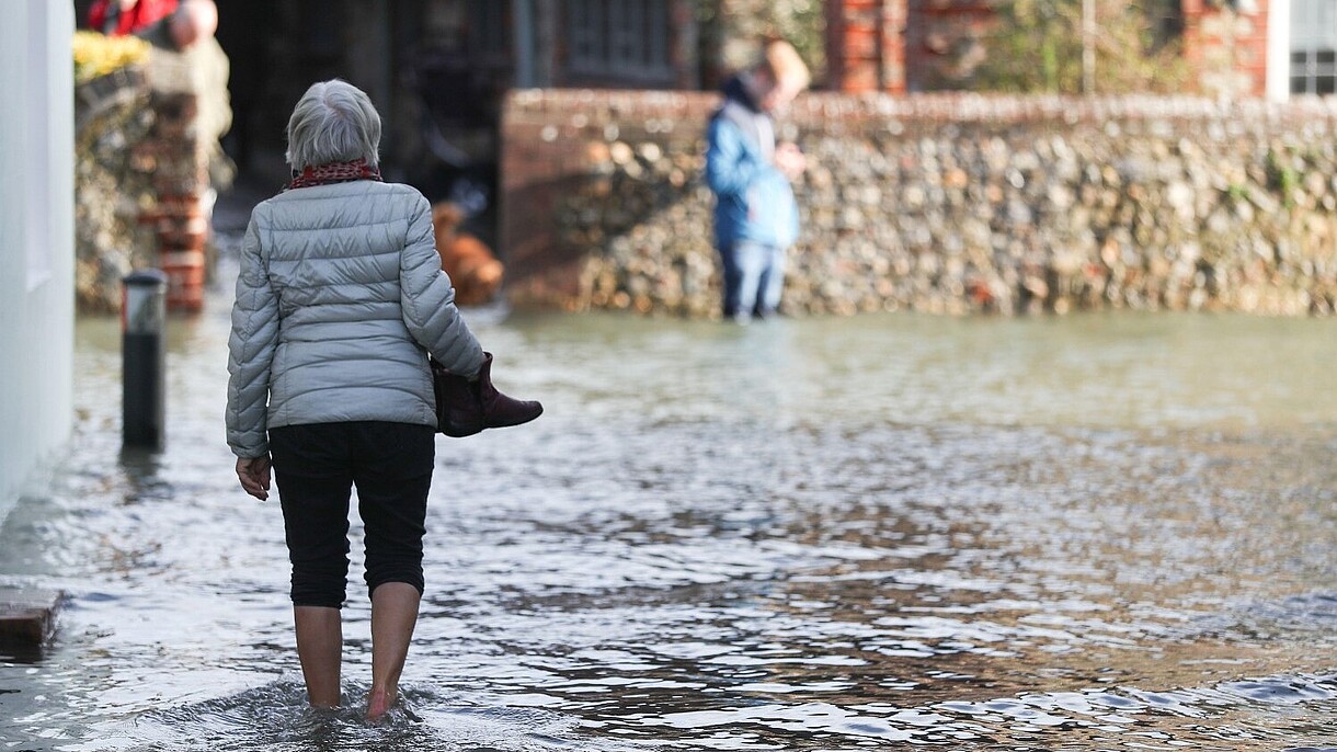 © PA Images / Alamy Stock Foto A person, photographed from the back, is walking barefoot through a flooded street. 