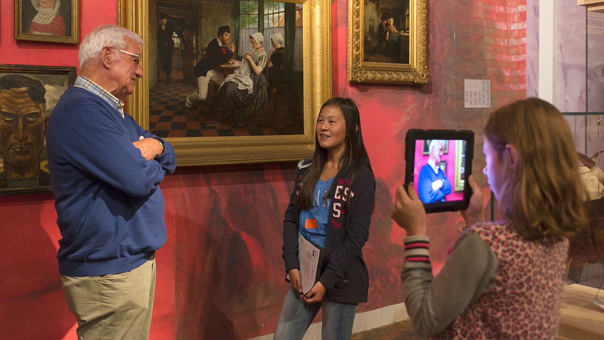 © Zeeuws Museum, Image: Anda van Riet One girl is using a tablet to film another girl who is talking with an older man, presumably about the painting behind them.