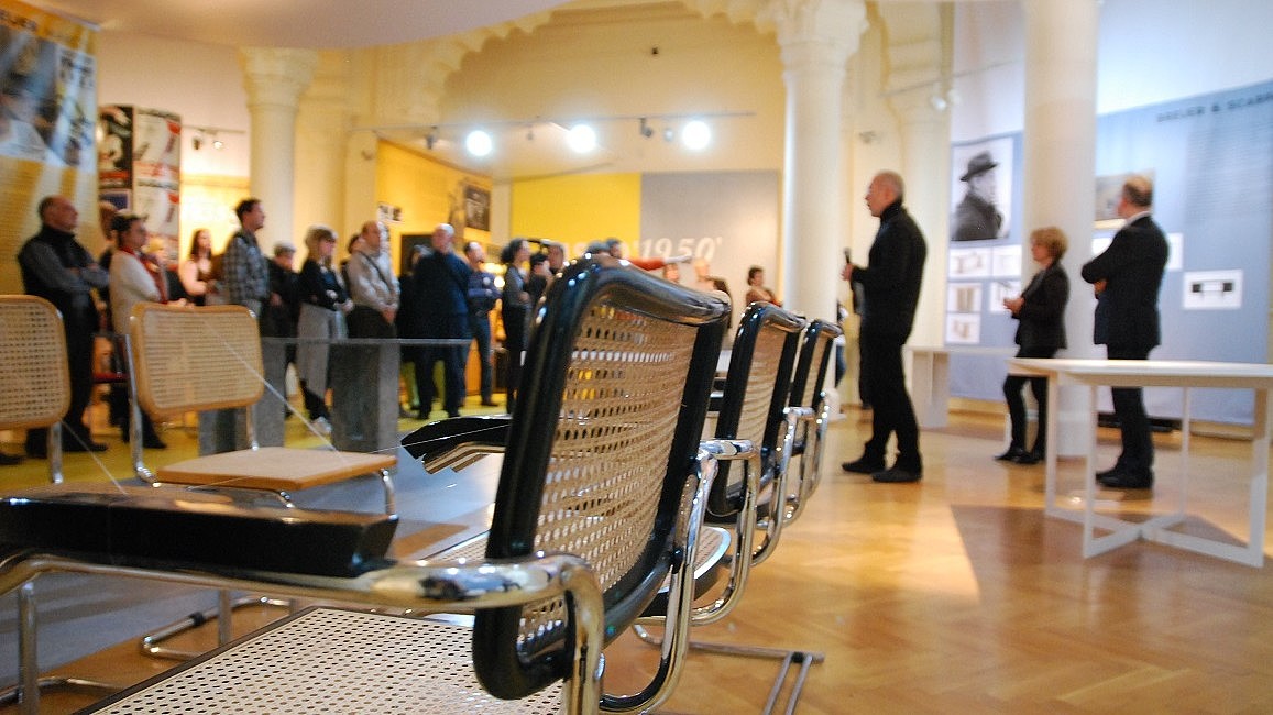  Two rows of chairs are facing each other in an exhibition space. In the background three people are giving a presentation to a group.