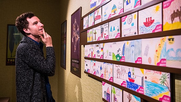  A man photographed from the side as he lightly touches his lips and smiles when admiring a wall full of what looks like paintings made by children.