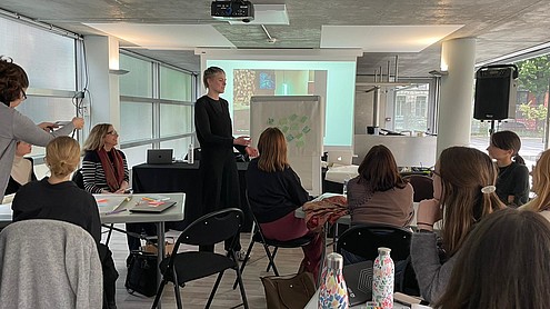 A person is standing next to a flipchart with sticky notes presenting something to a group of about 15 people seated around tables.   