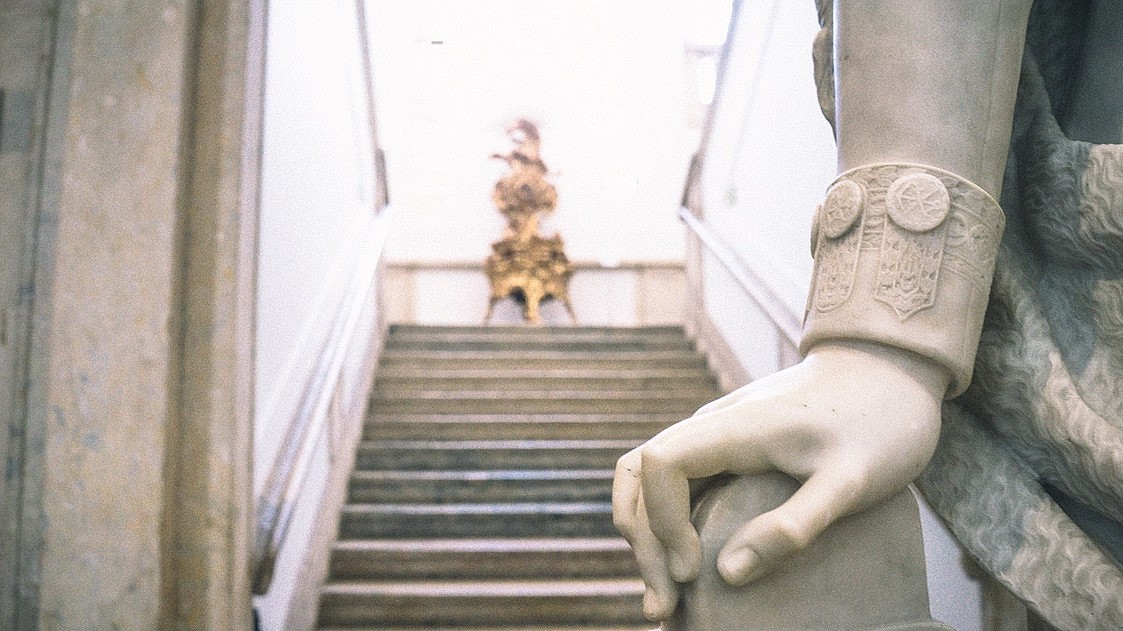 Detail of the hand of a marble statue. Behind a stone staircase is shown out of focus.