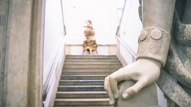  Detail of the hand of a marble statue. Behind a stone staircase is shown out of focus.