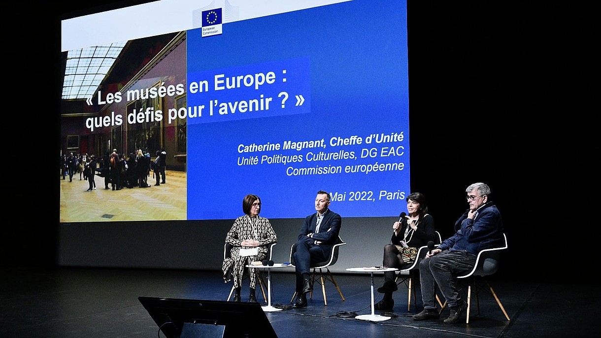  Four people sit on a lit stage. The rest of the room is dark. Behind them a presentation slide is projected. One person holds a microphone and talks.