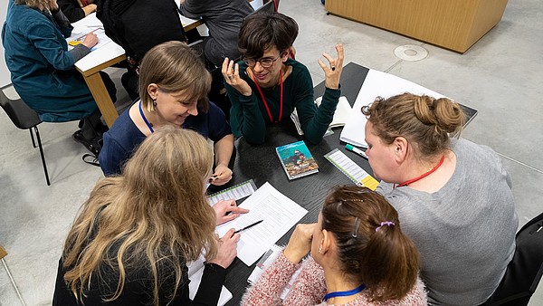 A group of people is sitting around a table looking at a piece of paper in their middle. One person is talking and gesturing with their hands.They are photographed from above.