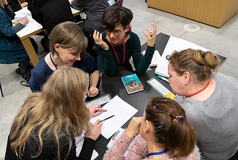 A group of people is sitting around a table looking at a piece of paper in their middle. One person is talking and gesturing with their hands.They are photographed from above.  