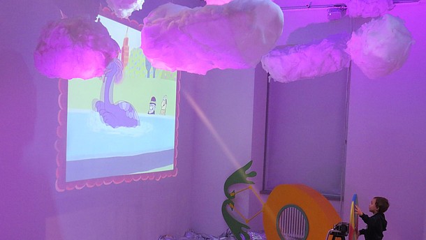  A boy is standing in a pink-lit room with cotton clouds hanging from the ceiling and silver foil laid out as water on the floor. He is steering a wheel whilst looking at a projector.
