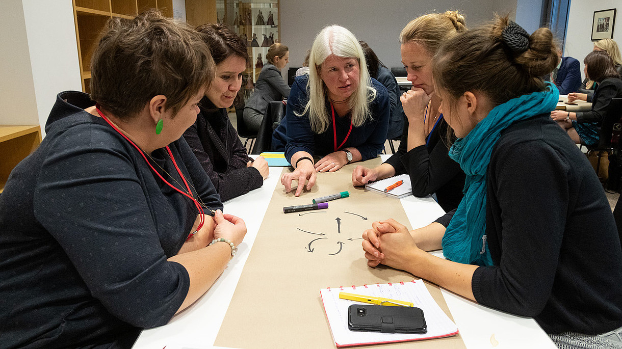 © Image: Berta Jänes © Image: Berta Jänes Someone leaning over a table is explaining something to a group of people sitting around it.