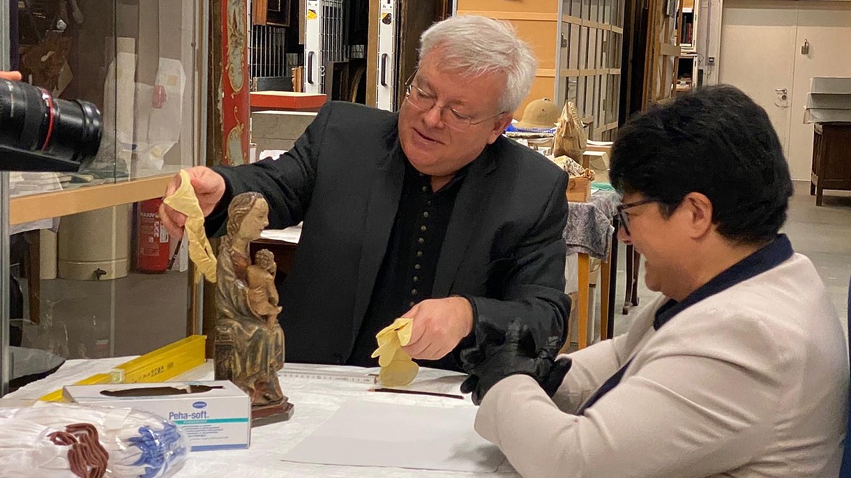  Sabine Verheyen looks at a small statue of a woman with a child on her lap, while another person says something and reaches out to the statue with a glove in hand, probably to carefully move it.