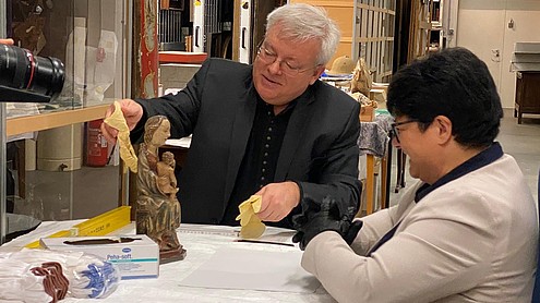 Sabine Verheyen looks at a small statue of a woman with a child on her lap, while another person says something and reaches out to the statue with a glove in hand, probably to carefully move it.  