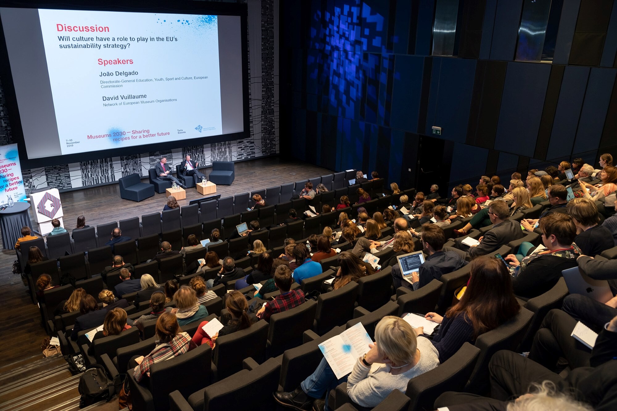 Auditorium full of people looking at two people on the stage. A sign shows that they are discussing EU culture policy.   © Image: Arp Karm