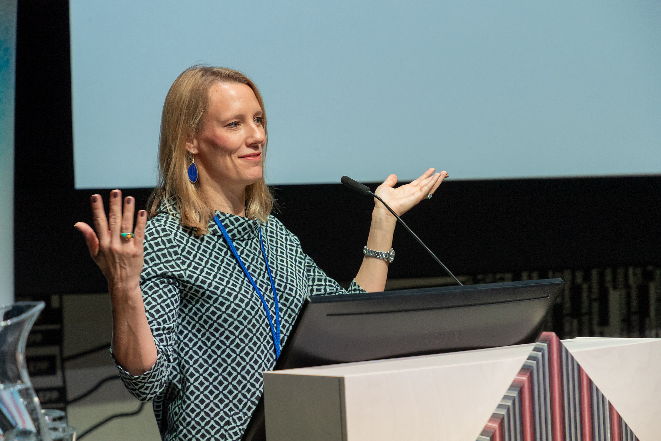 Woman standing behind a podium while giving a presentation is holding her hands up as if she is asking a question.  © Image: Arp Karm