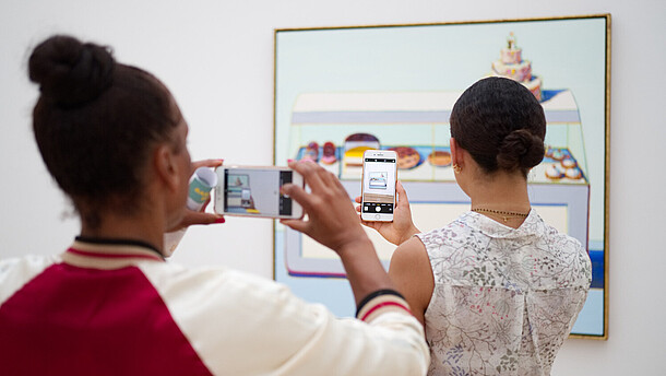  Two people have their backs to the camera as they both take a photo of a painting hanging on the wall. The person in the back is taking her photo of the painting through the phone screen of the person of the person standing in front of her.
