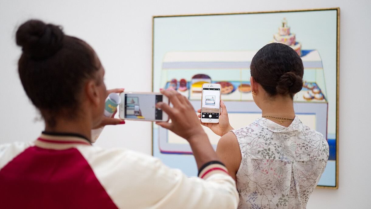 © Image: Christian Fregna © Image: Christian Fregna Two people have their backs to the camera as they both take a photo of a painting hanging on the wall. The person in the back is taking her photo of the painting through the phone screen of the person of the person standing in front of her.