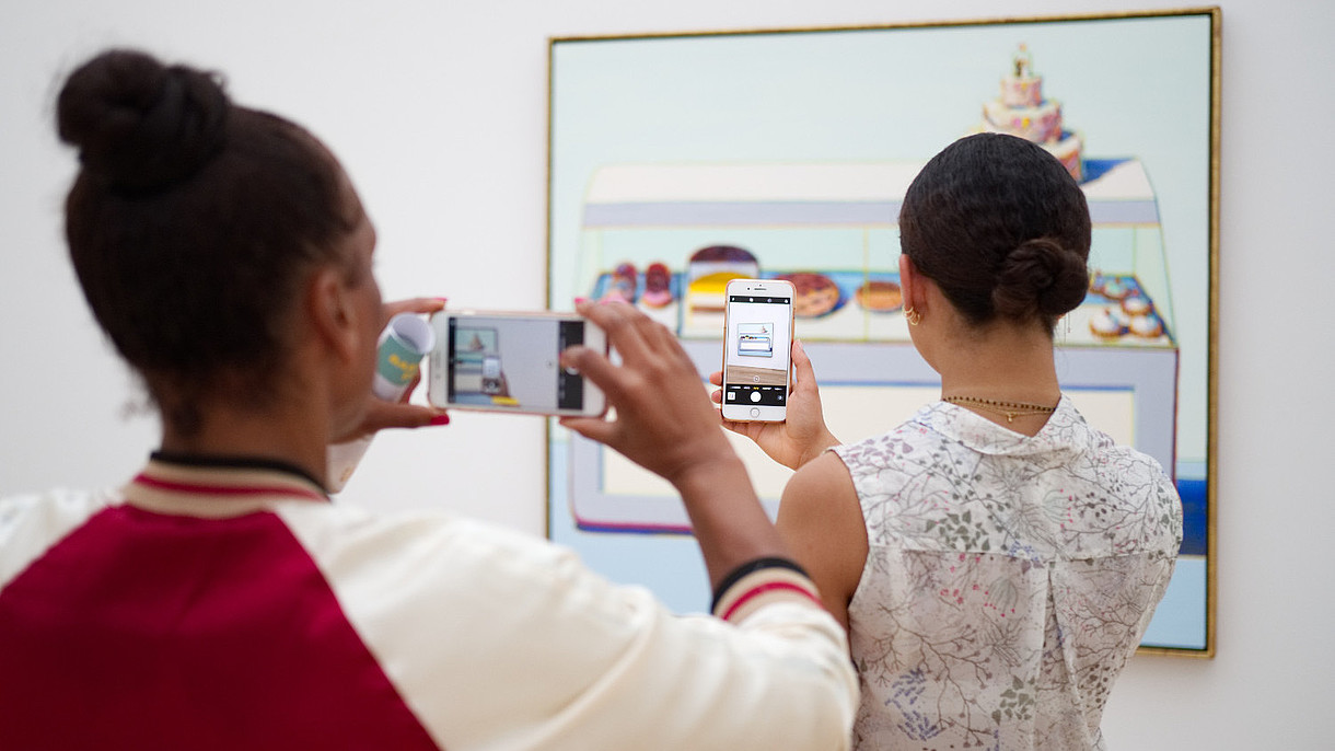 © Image: Christian Fregna Two people have their backs to the camera as they both take a photo of a painting hanging on the wall. The person in the back is taking her photo of the painting through the phone screen of the person of the person standing in front of her.