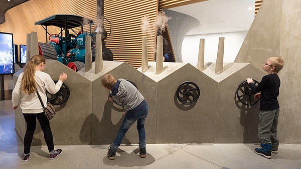 Three children are engaging with a museum display, by turning a steering wheel. In the background is a tractor.