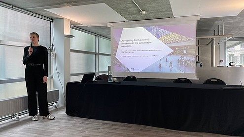 A person is standing next to a projector slide presenting with a handheld microphone. The slide reads “Advocating for the role of museums in the sustainable transition”.   