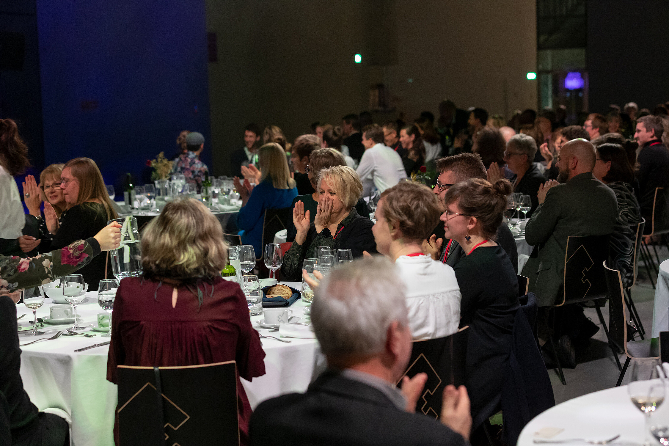 People are sitting down around round dinner tables. They are clapping their hand.   © Image: Arp Karm